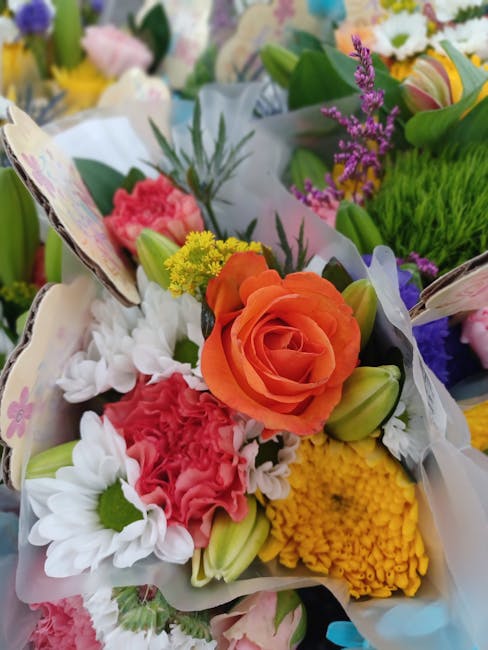 A vibrant floral bouquet featuring an orange rose as the focal point, surrounded by pink carnations, white daisies with green centers, yellow chrysanthemums, and small purple flowers. The flowers are 
