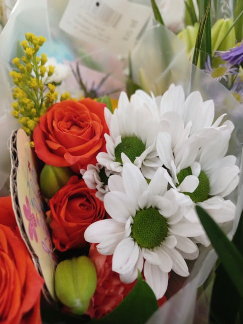 A vibrant floral bouquet featuring white daisies with green centers, paired with orange roses and small yellow filler flowers. The arrangement is wrapped in clear cellophane and positioned among other
