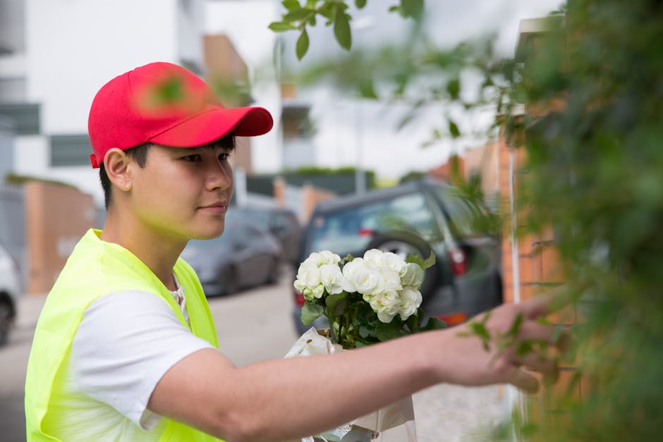 A young male florist with light skin, wearing a red baseball cap and a neon yellow vest, is seen arranging a bouquet of white roses and green foliage. He is standing outdoors on a residential street, 