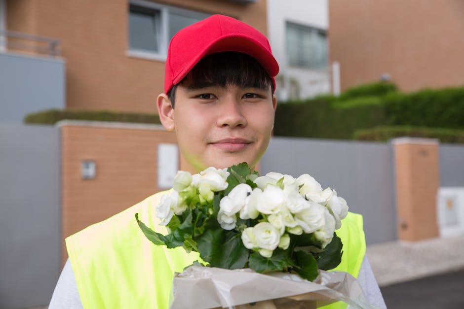 A young male delivery person wearing a red cap and a neon yellow vest holds a bouquet of fresh white roses wrapped in beige paper, standing outdoors in front of modern residential buildings with green