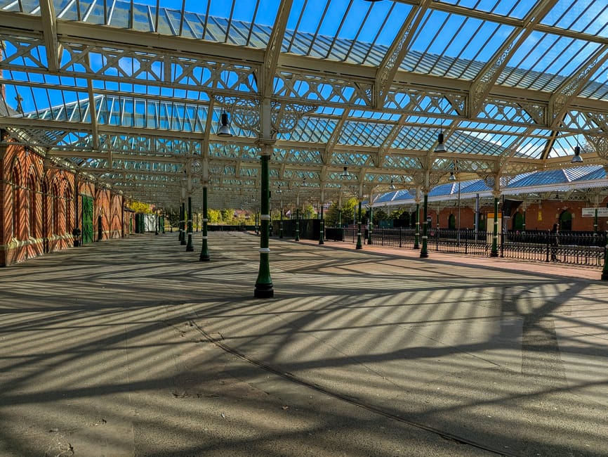 This image depicts an outdoor covered area at Hemel Hempstead station with a large glass and metal canopy casting intricate shadows on the paved ground. The structure features wrought iron decorative 