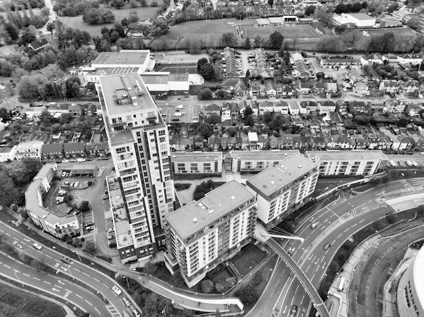Black and white aerial photograph of modern high-rise residential buildings situated above a multi-lane curved road, with surrounding smaller residential houses and green spaces visible in the backgro