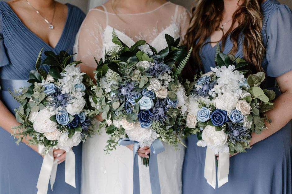 A close-up view of three women holding matching bridal bouquets featuring white and pale blue roses, white hydrangeas, and green foliage, with accents of blue thistle-like flowers and eucalyptus leave