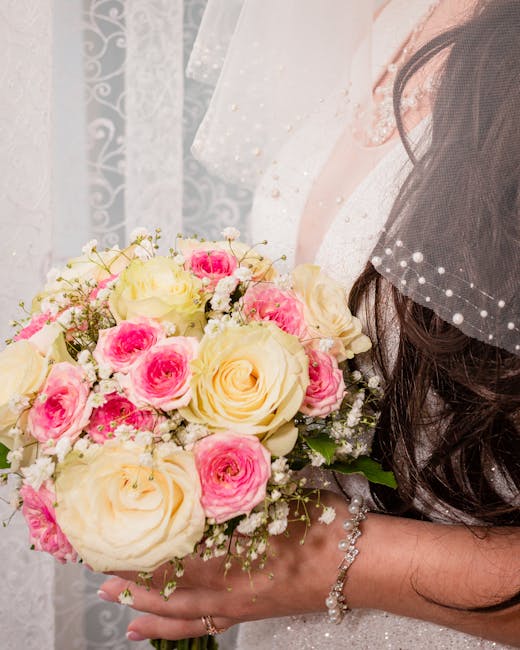 A bride's hands are gently holding a bouquet arrangement consisting of large, creamy white roses and smaller pink spray roses, accented by delicate baby's breath. The flowers appear fresh, with soft, 