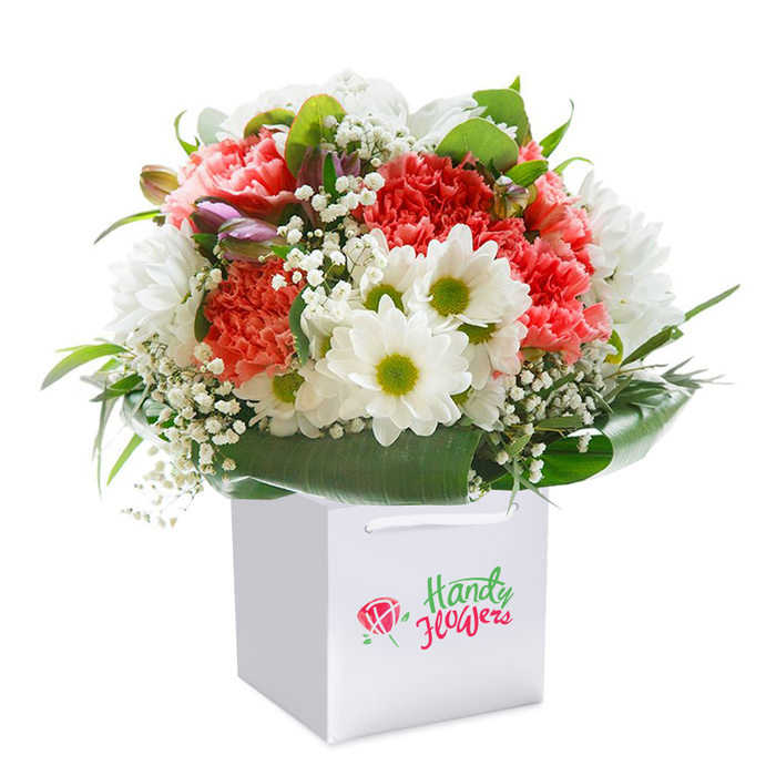 Echoing the lively heart of Hemel Hempstead, this beautifully arranged bouquet stands in a snowy white gift bag, its modern shape making it perfect for delivery along the bustling Marlowes or into the quiet avenues of Chaulden. The bouquet's coral and vibrant red carnations are distinguished by layers of feathery, ruffled petals, creating waves of rich texture and warmth. Among these, white daisies with radiant yellow-green centres cluster happily, reminiscent of the wildflowers glimpsed on walks through Gadebridge Park. Baby's breath is scattered throughout the blooms, its tiny white blossoms weaving a veil of airiness, while a few slender purple-tinted buds peep from the left, lending subtle depth. Glossy deep green foliage wraps the bouquet in a natural embrace, giving structure and form, and evoking the lush hedgerows lining local trails. The white bag, finished with sturdy ribbon handles, speaks to the practical needs of everyday delivery across Hemel Hempstead. The entire floral display is captured in a professional photograph on an immaculate white backdrop, ensuring each detail glows with clarity. This arrangement, with its sophisticated balance and vibrant freshness, is suited for birthdays, anniversaries, or heartfelt everyday gestures, all reflecting the trusted skill and care of Hemel Hempstead's local florists.